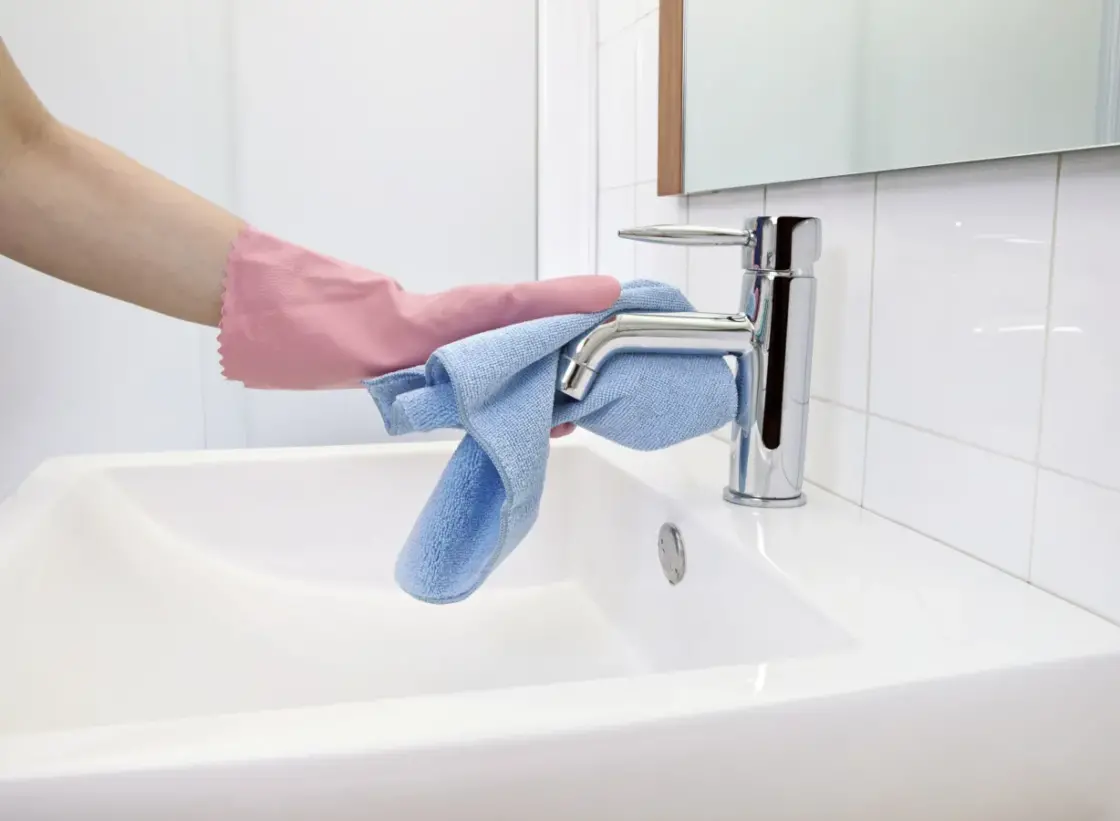 Woman cleaning hard water stain off the faucet with microfiber cloth and gloves