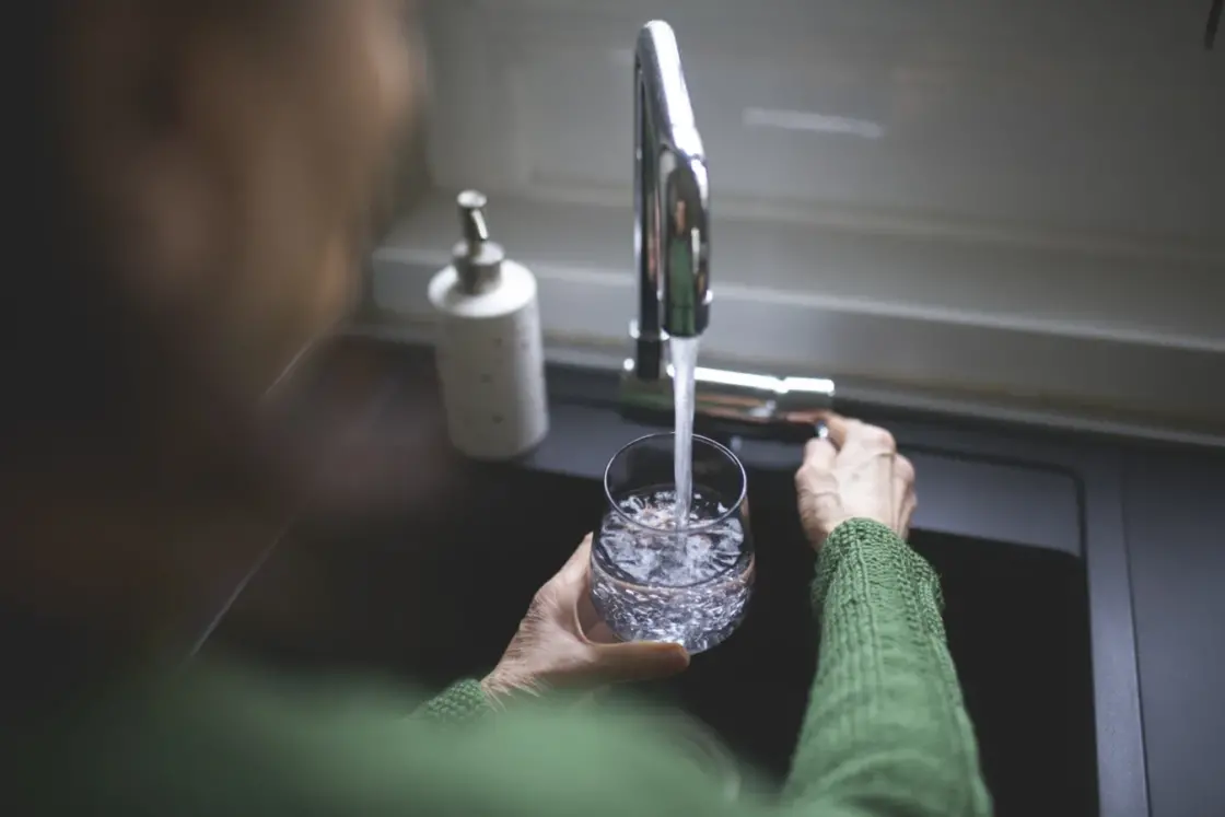 Close up of a senior woman's hand filling a glass of filtered water from the kitchen sink tap. Cost of a water softener